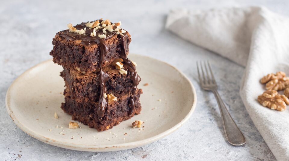 Una golosa torretta di tre quadrotti di brownies al cioccolato, ricoperti da una colata di cioccolato fuso e granella di noci, appoggiata su un piatto in ceramica chiara.
