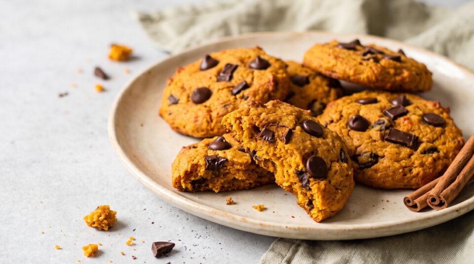 Un piatto in ceramica con dei cookies zucca e gocce di cioccolato appena sfornati, appoggiati su un tavolo di legno rustico. Un biscotto è spezzato a metà e mostra l'interno morbido con il cioccolato fondente fuso. Sfondo luminoso e pulito.