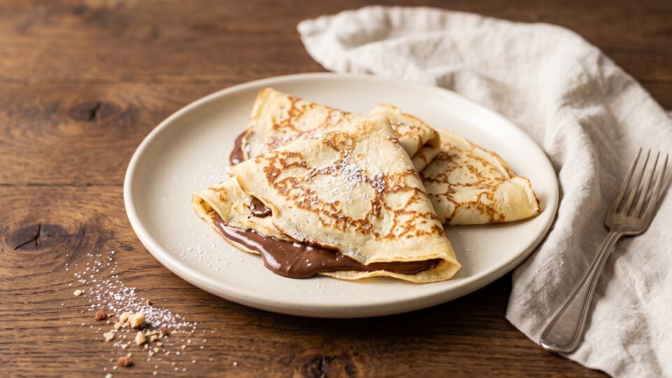 crêpes sottilissime e morbide piegate a triangolo, farcite con abbondante Nutella fusa che cola dai bordi. Le crêpes sono spolverate di zucchero a velo e appoggiate su un tavolo di legno rustico luminoso.