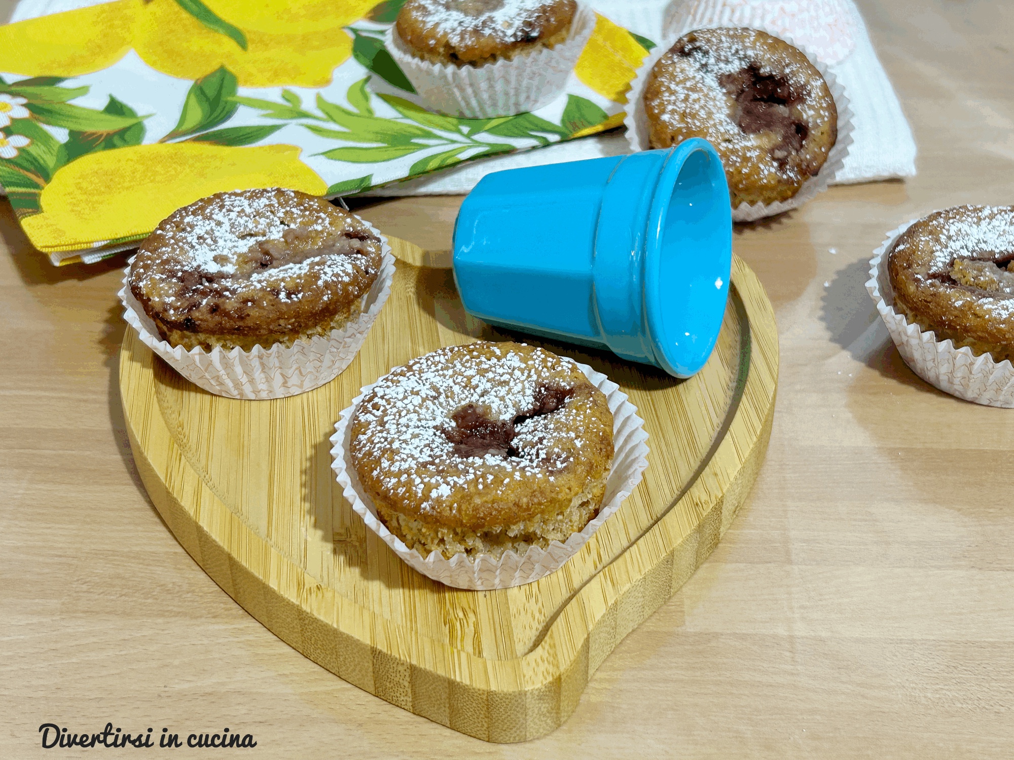Quattro muffin integrali con cuore di marmellata scura e una spolverata di zucchero a velo, disposti su un vassoio di legno a forma di cuore con accanto una tazzina azzurra e un tovagliolo con limoni