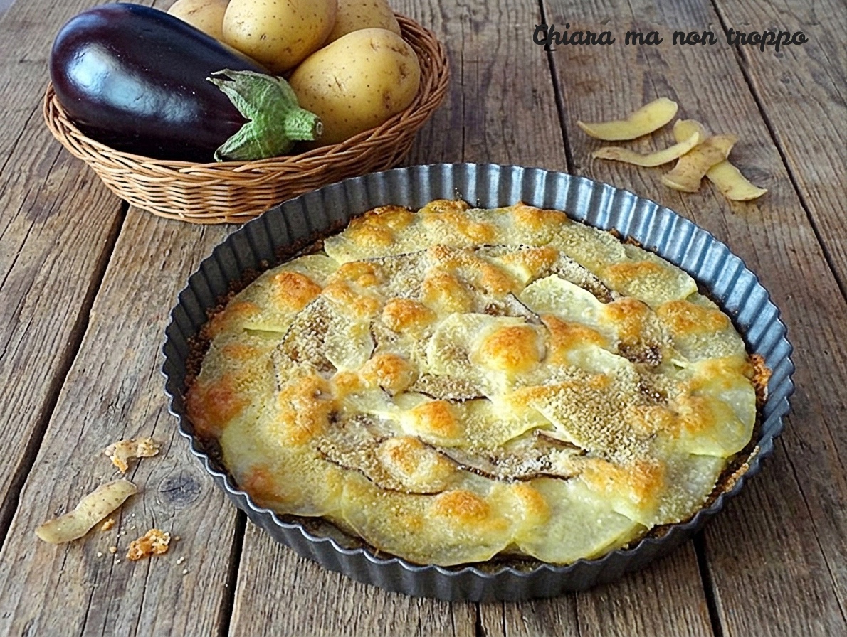Una vista dell'alto inquadra un tortino di patate e melanzane gratinato, servito in una tortiera di alluminio con i bordi scanalati. Il tortino è ricoperto da uno strato di parmigiano e pangrattato dorato. Sullo sfondo, un cesto di vimini con melanzane e patate intere
