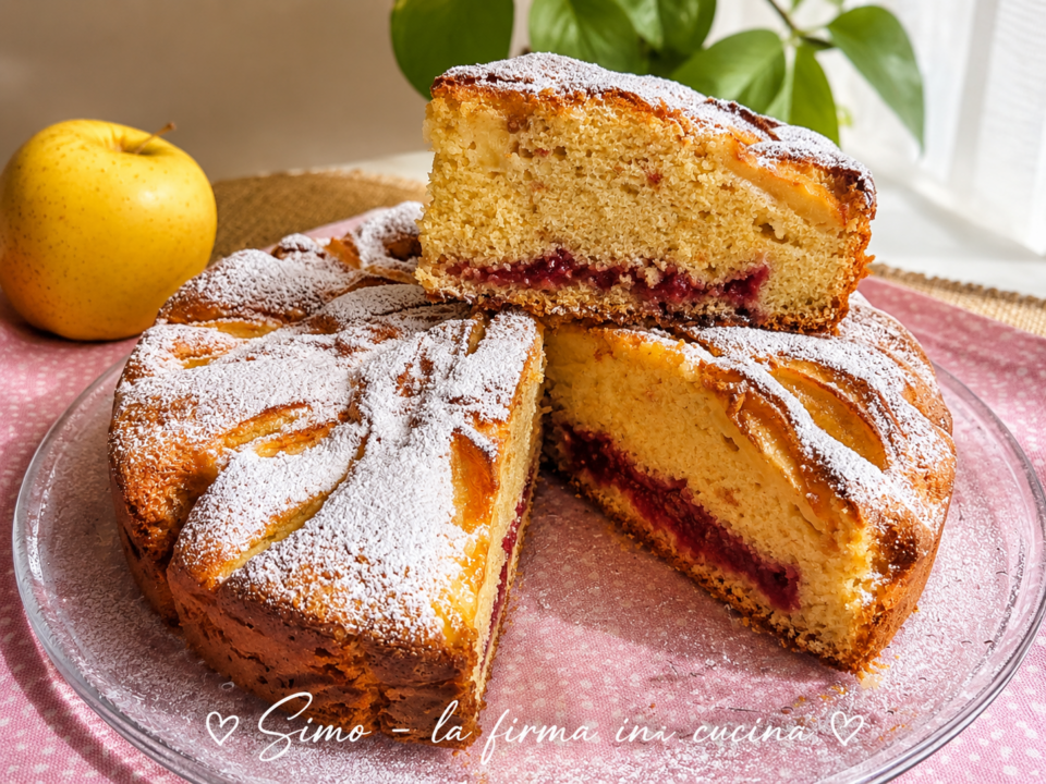 Torta di mele soffice con marmellata di fragole, con fetta tagliata decorata con lo zucchero a velo servita sul piatto di vetro , alle spalle una mela decorativa 