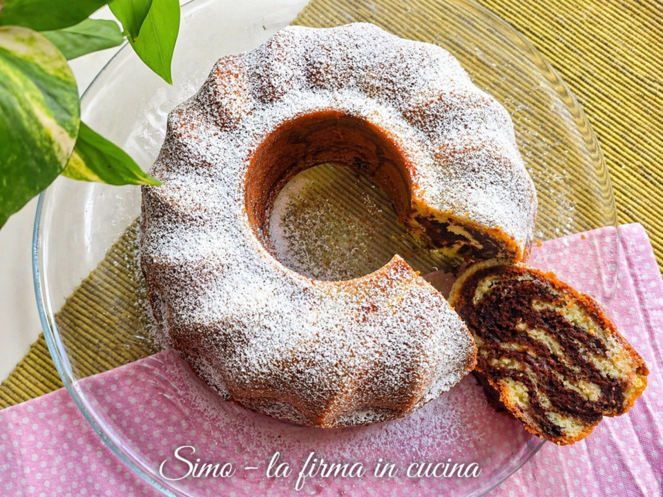 Ciambella marmorizzata allo yogurt con zucchero a velo e fetta tagliata che mostra l'effetto marmorizzato al cacao, servita su un piatto di vetro con tovaglietta decorativa.